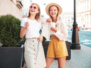 Two young beautiful smiling hipster female in trendy summer clothes.Sexy carefree women posing in the street. Positive pure models having fun at sunset. They drinking coffee or tea in plastic cup