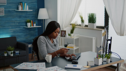 Black woman holding electronic credit card typing online payment on computer keyboard while ordering electronic tablet during online sale. Customer young woman sitting at desk table in living room
