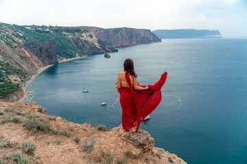 Fototapeta na wymiar A girl with loose hair in a long red dress descends the stairs between the yellow rocks overlooking the sea. A rock can be seen in the sea. Sunny path on the sea from the rising sun