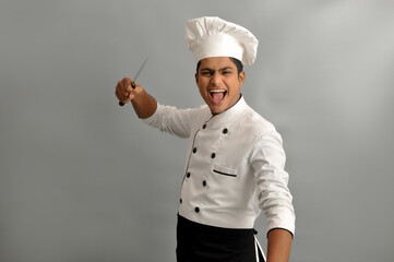 Indian chef smiling and giving Action pose with his knives while standing over grey background