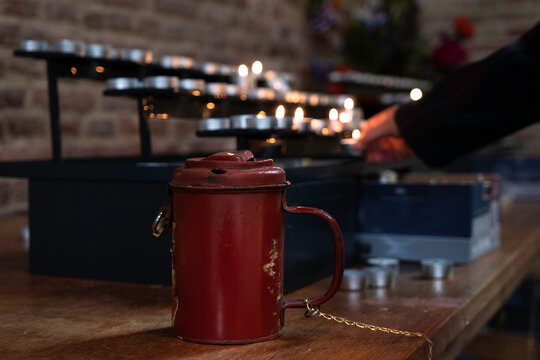 Woman Lights Candle In Church After Throwing Money Into Red Metal Collection Box For Donations. Focus On The Collection Box On The Table