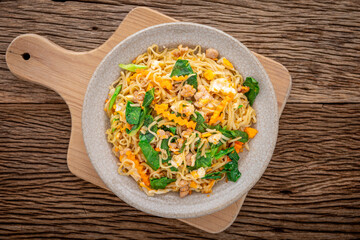 stir fried instant noodles with pork, egg, chinese kale, carrot and pepper in crazing ceramic plate on cutting board on rustic natural wood texture background with copy space for text, top view