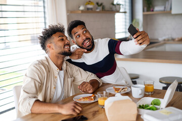 Portrait of young adult brothers in kitchen indoors at home, taking selfie.