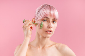 Portrait of young naked woman with pink hair using natural jade facial roller for skin care, posing...