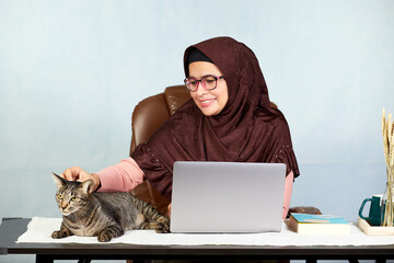 Asian Muslim woman smiling working on laptop computer and her hand patting a cat on the desk