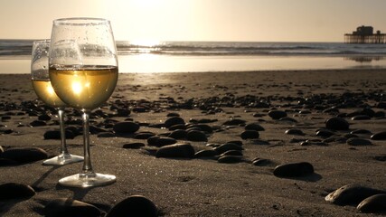Two wineglasses on sandy ocean beach. Couple of glasses and pebbles, white wine for romantic date near sea water. Cozy beachfront sunset near tide waves. Seamless looped cinemagraph. California, USA.