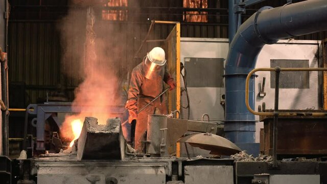 A foundry worker wearing full heat-protective clothing does the dangerous job of stoking the furnace of molten metal causing orange sparks and smoke to fly.
