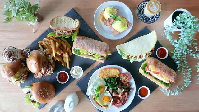 Cafe latte pushed into frame next to brunch flat lay with sandwiches, burger and chips.