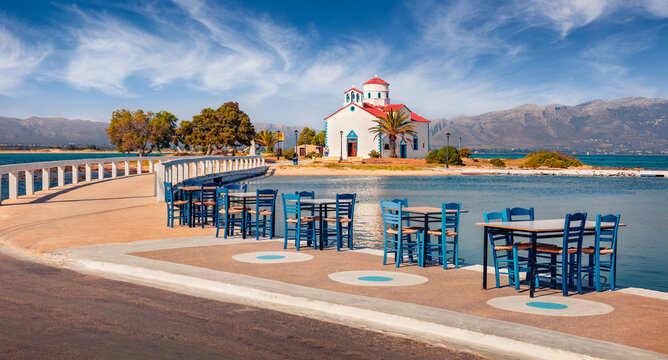 Romantic summer view of of Saint Spiridon, Greek Orthodox church and empty tables of smal coastal resraurant. Beautiful outdoor scene of Elafonisos island, Greece. Bright Mrditerranean seascape.