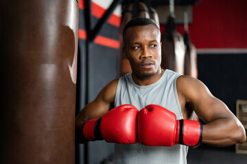 Young African American muscular man in a white tank top wearing a red boxing gloves standing in front of a punching bag - Powered by Adobe