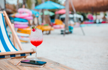 Close-up, juice with red cherry in champagne glass and mobile phone, book on wooden table on sand beach. Concept relaxing after work on summer