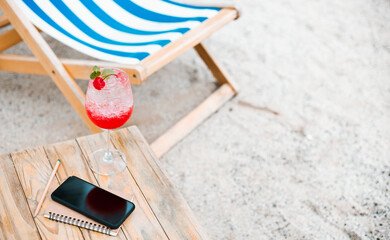 Close-up, juice with red cherry in champagne glass and mobile phone, book on wooden table on sand...