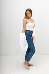 young attractive caucasian woman with long brown hair in corset, shirt, blue jeans on white studio background. skinny pretty lady posing with bare feet at bright room. portrait of beautiful female
