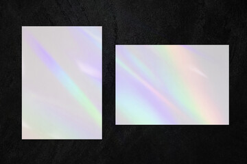 Two empty white vertical and horizontal rectangle a4 poster, business card or flyer mockups with overlay of rainbow light refraction caustic effect and shadow on black concrete background.