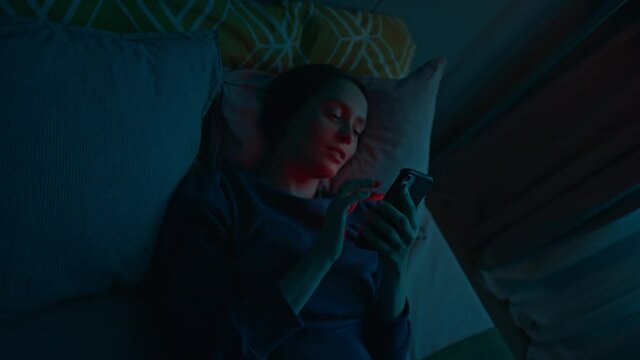 Top view of young smiling woman lying on bed alone in dark bedroom and using her smartphone for online communication, surfing and chatting. Modern technology concept. Slow motion cinematic shot