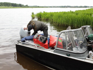 Boater man repairs the 50 hp four stroke outboard motor on transom of boat near shore grass, repair...