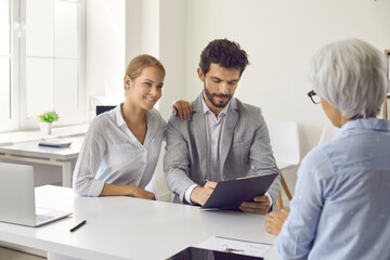 Smiling married millennial couple signing agreement document during meeting in bank office. Two happy young clients putting signatures on contract paper given by mortgage agent, realtor or loan broker