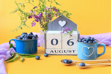 Calendar for August 6 :the name of the month of August in English, cubes with the numbers 0 and 6, blueberries in blue cups, bouquets of flowers, yellow background, side view
