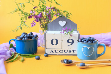 Calendar for August 9 :the name of the month of August in English, cubes with the numbers 0 and 9, blueberries in blue cups, bouquets of flowers, yellow background, side view