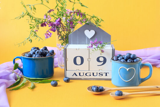 Calendar for August 9 :the name of the month of August in English, cubes with the numbers 0 and 9, blueberries in blue cups, bouquets of flowers, yellow background, side view