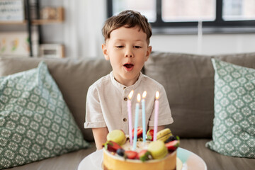 holidays, celebration and people concept - happy little boy blowing candles on birthday cake at home party