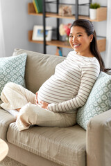 Obraz premium pregnancy, rest, people and expectation concept - happy smiling pregnant asian woman sitting on sofa at home