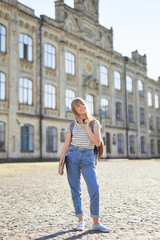 Attractive happy young blonde high school girl or college student with backpack and laptop wearing jeans standing in university campus. Educational or studying concept. High quality vertical image