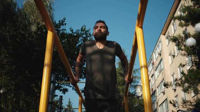 Caucasian man with beard and dreadlocks does push ups on parallel bars outside. Sports activities on outdoor ground and healthy lifestyle. Fitness and strength exercises for muscles of arms.