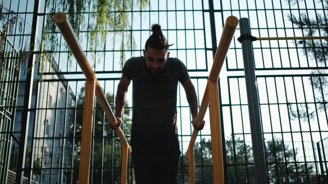 Caucasian man with beard and dreadlocks does push ups on parallel bars outside. Sports activities on outdoor ground and healthy lifestyle. Fitness and strength exercises for muscles of arms.