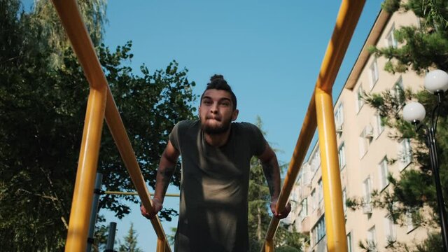 Caucasian man with beard and dreadlocks does push ups on parallel bars outside. Sports activities on outdoor ground and healthy lifestyle. Fitness and strength exercises for muscles of arms.