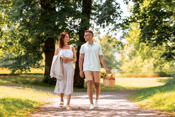 Fototapeta premium leisure, vacation and people concept - happy couple with food in picnic basket and blanket walking at summer park