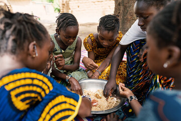 Group of coulorfully dressed black girls sitting around a big metal bowl, sharing a typical African meal made out of rice and vegetables
