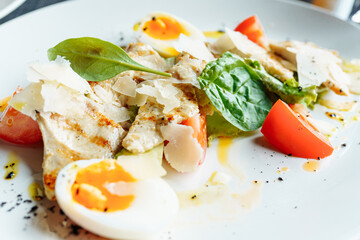 Classical Caesar salad with roasted chiken, eggs, lettuce leaves, parmesan and cherry tomatoes on the white plate close up