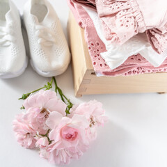 A stack of pink baby girls clothes in a wooden box, white sneakers. Toddlers set of clothes n white background with copy space, decorated with pink flowers