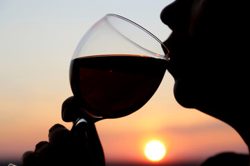 Naklejka na ściany i meble Woman drinking red wine, silhouette of face with glass at sunset. Enjoying alcohol, romantic dinner at the resort, luxury life