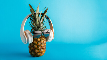 Creative funny pineapple face wearing sunglasses headphones. Pineapple face levitating listening to music on color blue summer background. Long web banner with copy space.