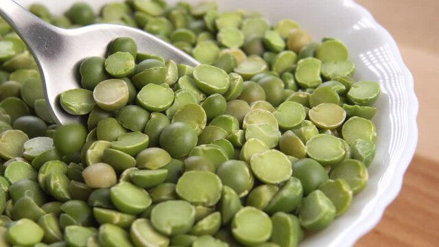 Metal spoon stirring green split dry peas in a full white bowl on a wooden rustic table. Macro shot