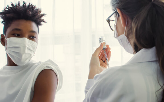 African Teen Young Man Wearing Face Mask, Getting Vaccination To Protect Or Prevent Virus, Getting Scary Of Vaccine Injection While Female Doctor Preparing Syringe To Vaccinate Patient At Hospital.