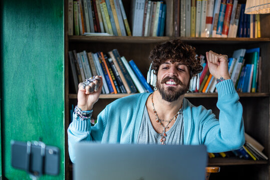 Successful influencer who listens to music and exults victoriously in a video call, young man with tattoos and piercings who creates content online with laptop