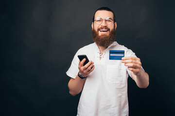 A nice picture of a young bearded man smiling at the camera and holding a phone and a credit card ready to go shopping online