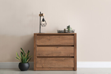 Wooden chest of drawers with houseplants and lamp near beige wall in room - obrazy, fototapety, plakaty