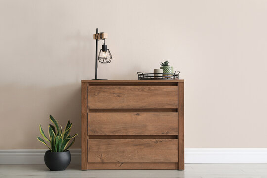 Wooden chest of drawers with houseplants and lamp near beige wall in room