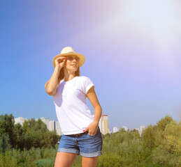 Young woman in a hat and sunglasses against a background of green foliage, houses and blue sky. The sun shines brightly