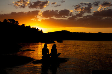 Naklejka na ściany i meble Black silhouette of two friends sitting on a lake in the golden sunset. Copy space.