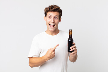 young handsome man looking excited and surprised pointing to the side and holding a beer bottle