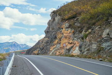 Colorful autumn landscape with mountain highway along rocks in sunshine. Bright alpine scenery with mountain road in autumn colors. Highway in mountains in fall time. Road along beautiful rocky wall.