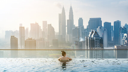 Fototapeta premium young Asian woman enjoying in hotel roof top swimming pool, city view with blue sky for vacation travel, lifestyle of beautiful tourist girl with luxury outdoor skyscraper, Kuala Lumpur, Malaysia