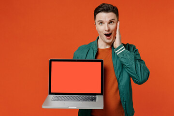 Shocked fun young brunet man 20s wear red t-shirt green jacket hold use laptop pc computer with blank screen workspace area keeping mouth wide open isolated on plain orange background studio portrait