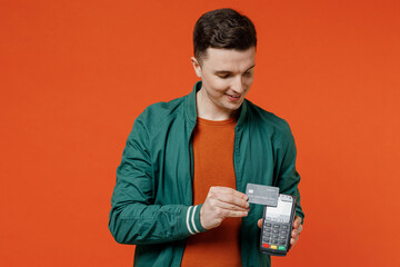 Satisfied fun young brunet man 20s wears red t-shirt green jacket use wireless modern bank payment terminal to process acquire credit card payments isolated on plain orange background studio portrait