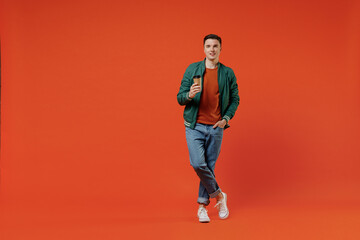 Fototapeta na wymiar Full size body length smiling happy young brunet man 20s wears red t-shirt green jacket hold takeaway delivery craft paper brown cup coffee to go isolated on plain orange background studio portrait
